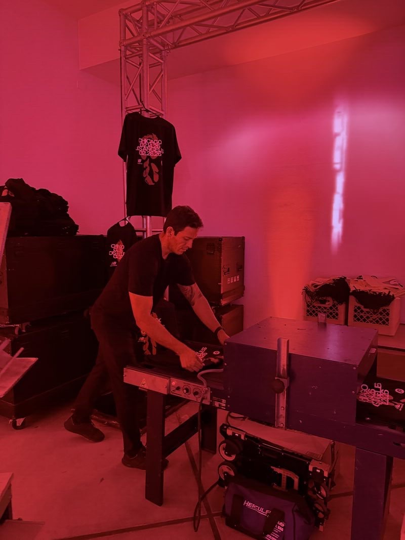 Crew member feeding freshly printed shirts through a conveyor dryer in a red-lit backstage production area with road cases