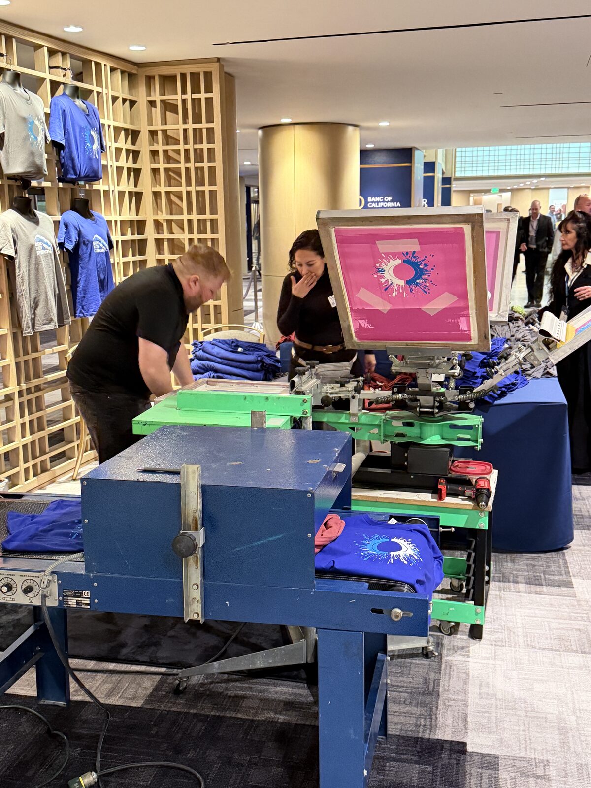 Two crew members running a screen printing press beside a conveyor dryer with royal blue shirts at a corporate summit