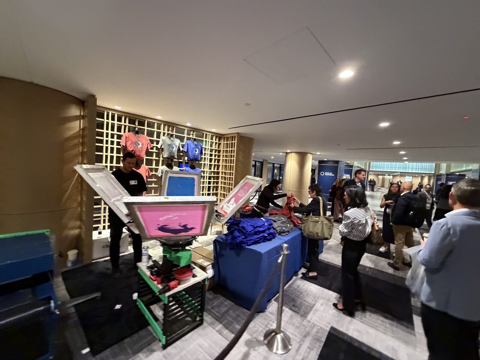 Live screen printing booth with a merch display wall and stacked shirt table drawing attendees in a convention hall