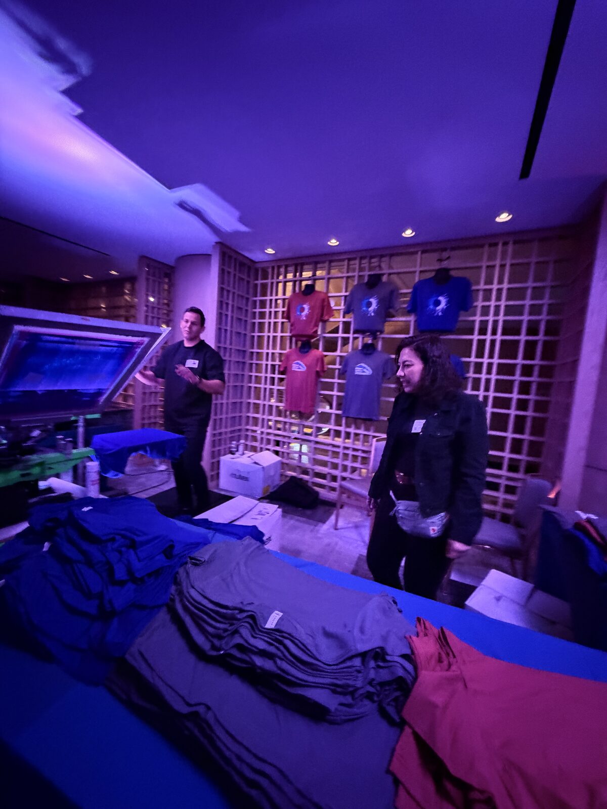 Crew member walking a guest through stacks of gray, navy, and red blanks on a garment table under purple uplighting
