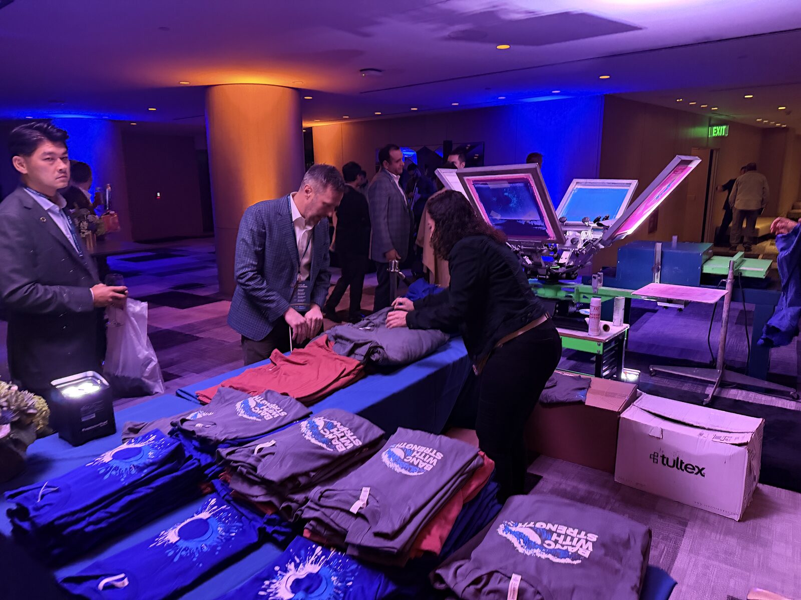 Executives in suits browsing folded tees at a long blue garment table beside a screen printing press with pink-lit screens