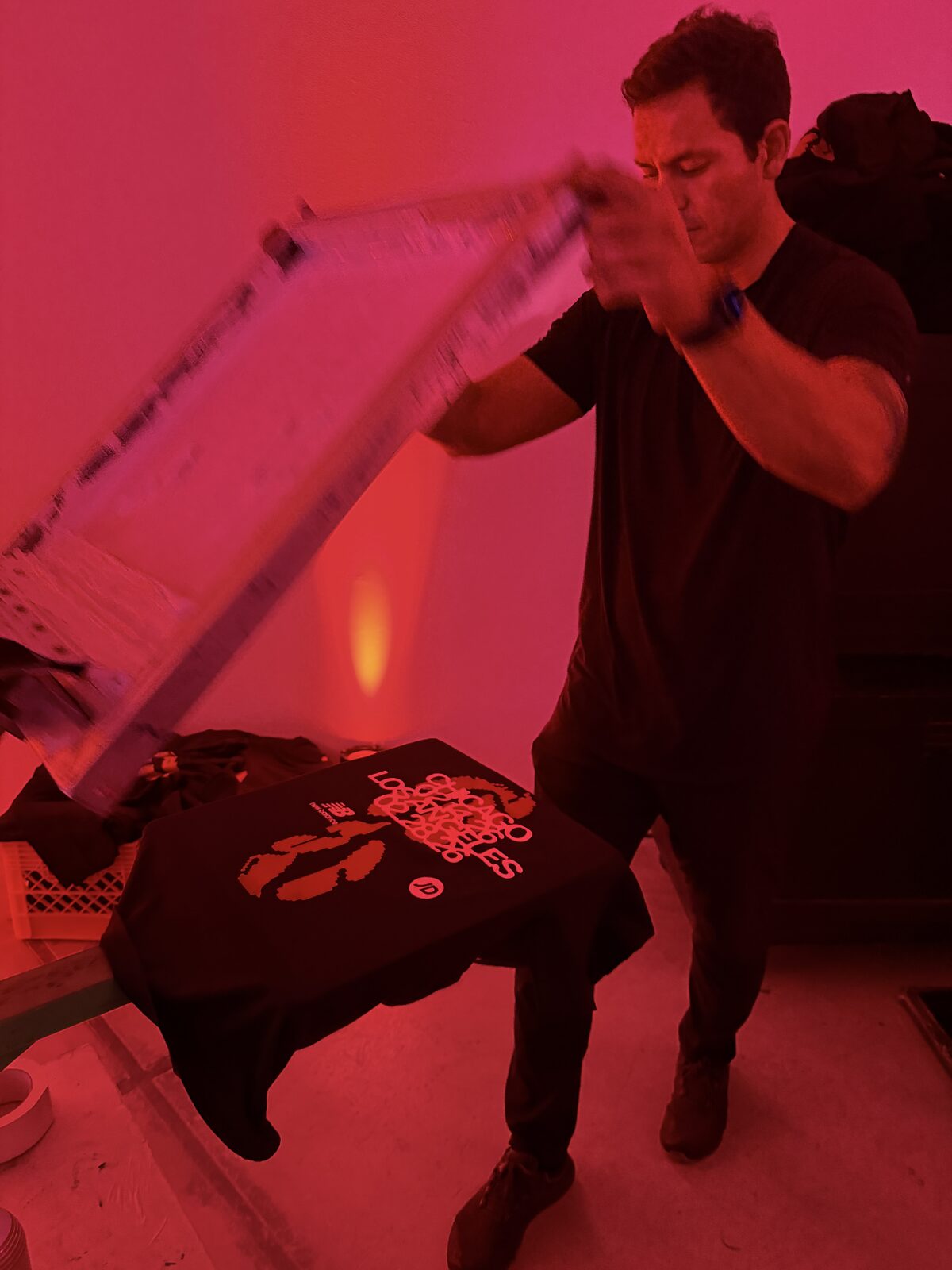 Printer lifting a screen off a freshly printed black launch tee under deep red light