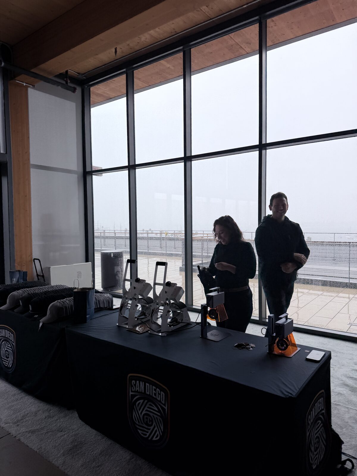 Two crew silhouetted at a San Diego FC table of cap heat presses in front of floor-to-ceiling windows