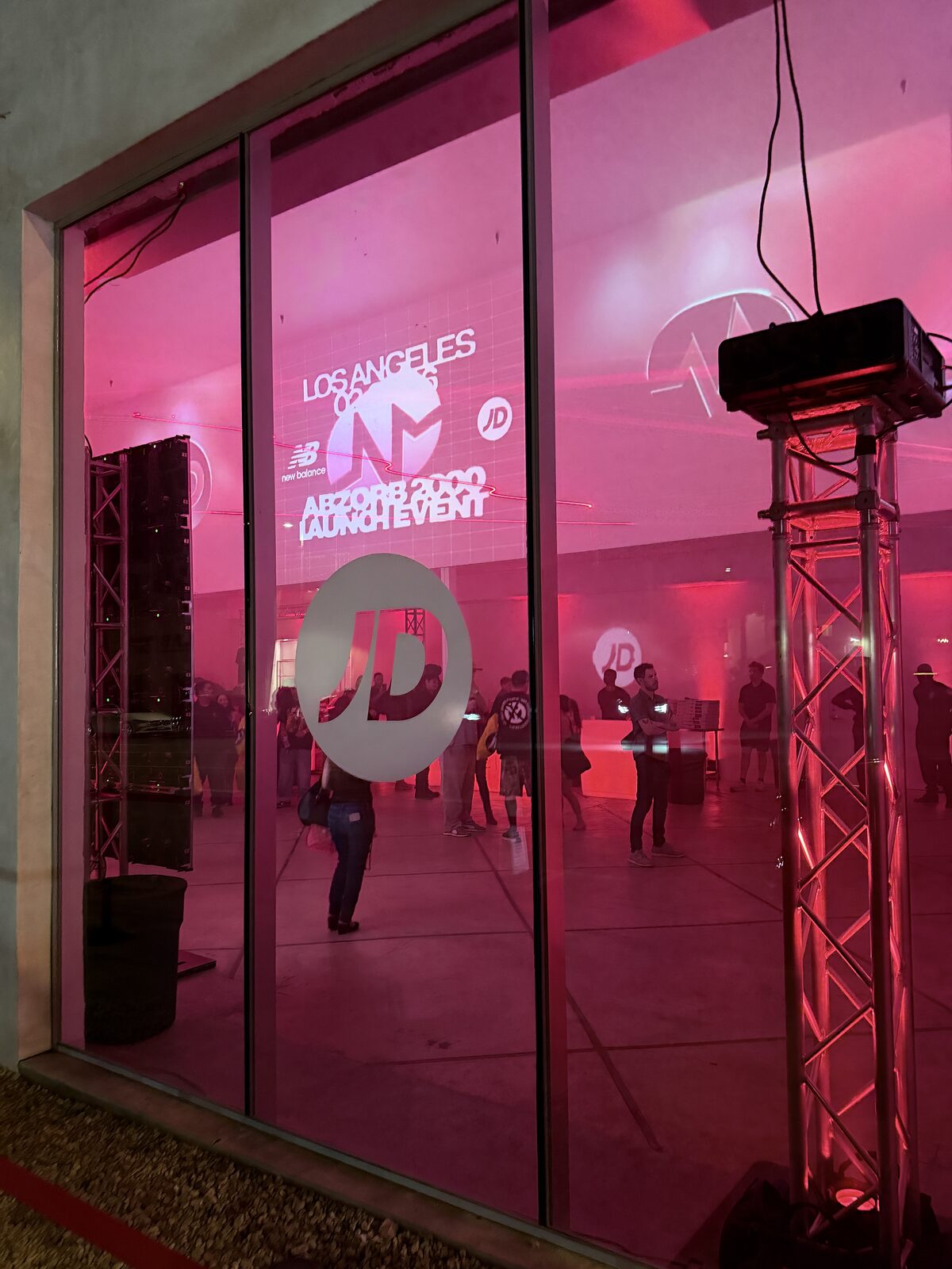 Pink-lit launch venue seen through glass with a Los Angeles projection wall and a lighting truss outside the window
