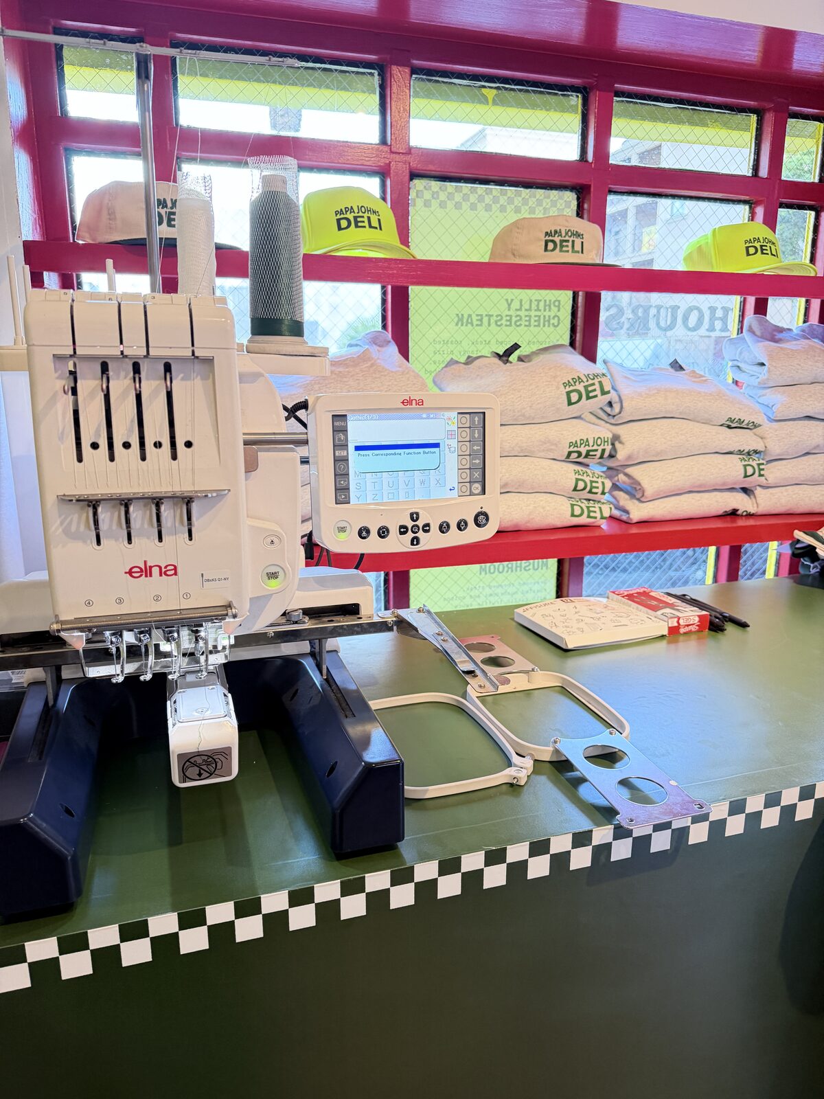 Multi-needle embroidery machine running on a green counter with neon yellow caps and cream crewnecks stacked behind it