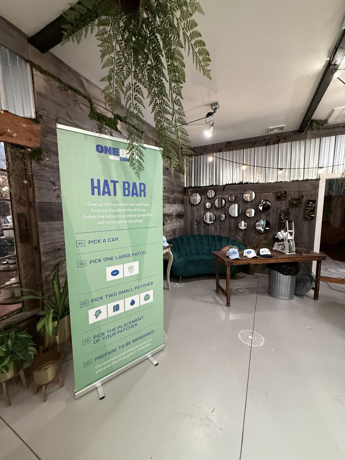 Lounge-style hat bar with a step-by-step instruction banner, teal velvet sofa, mirror wall, and caps staged on a wood table