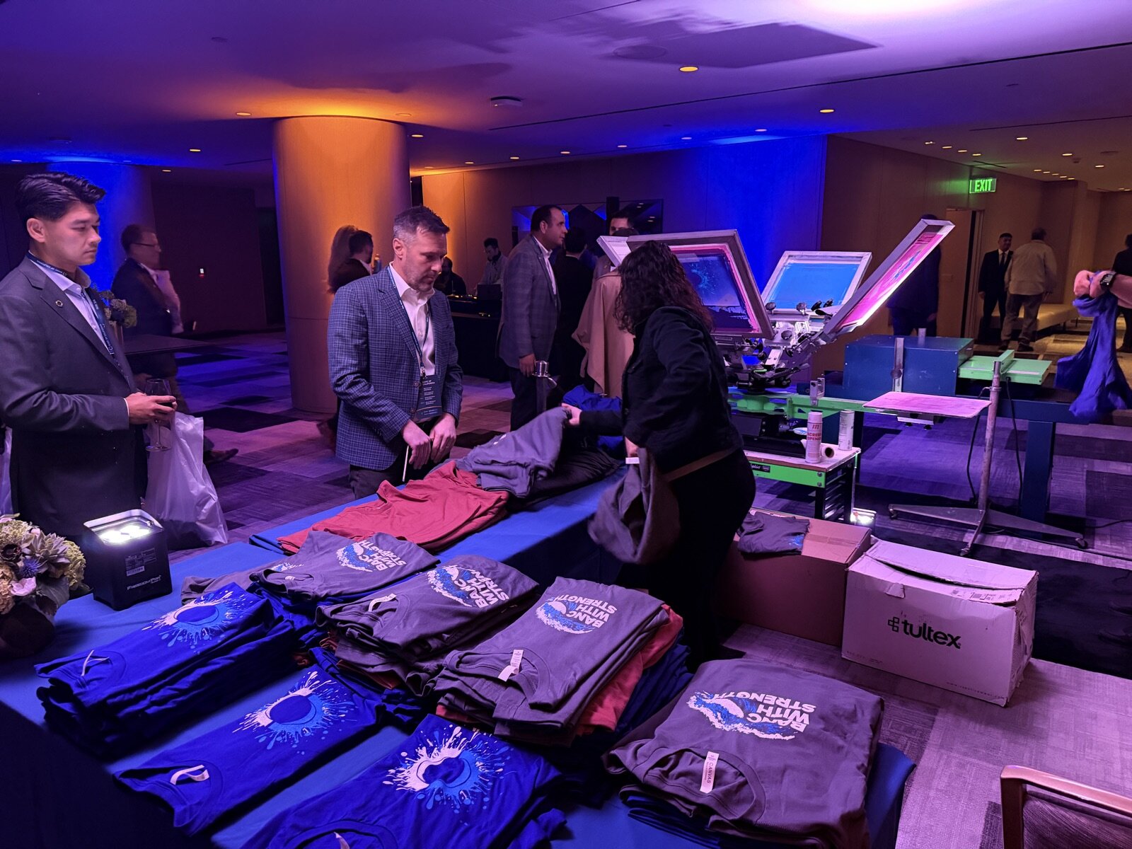 Merch Troop crew running two screen printing presses beside long tables of folded shirts at a corporate event