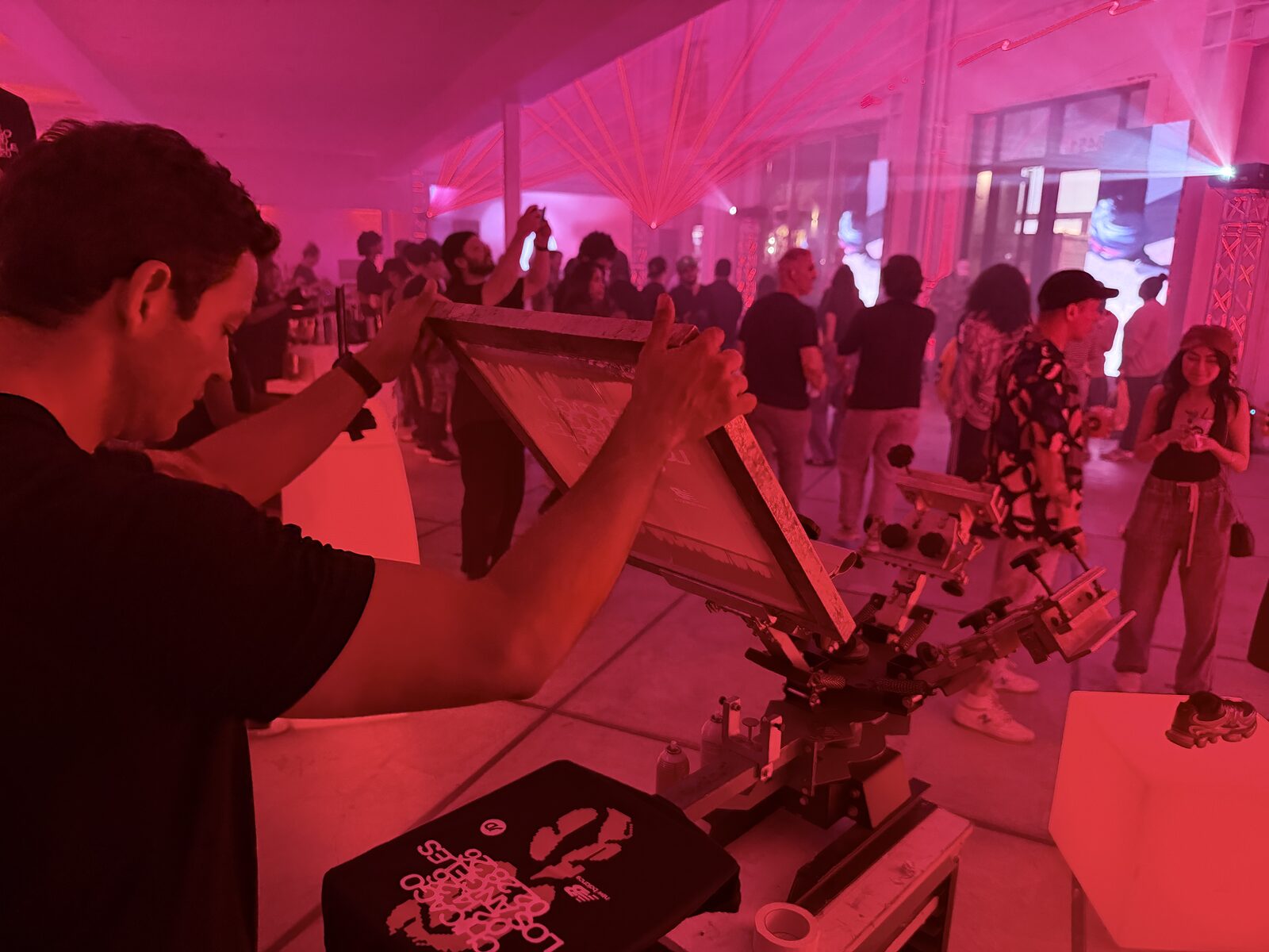 Screen printer raising a frame over a New Balance shirt while launch party guests film the press under pink light