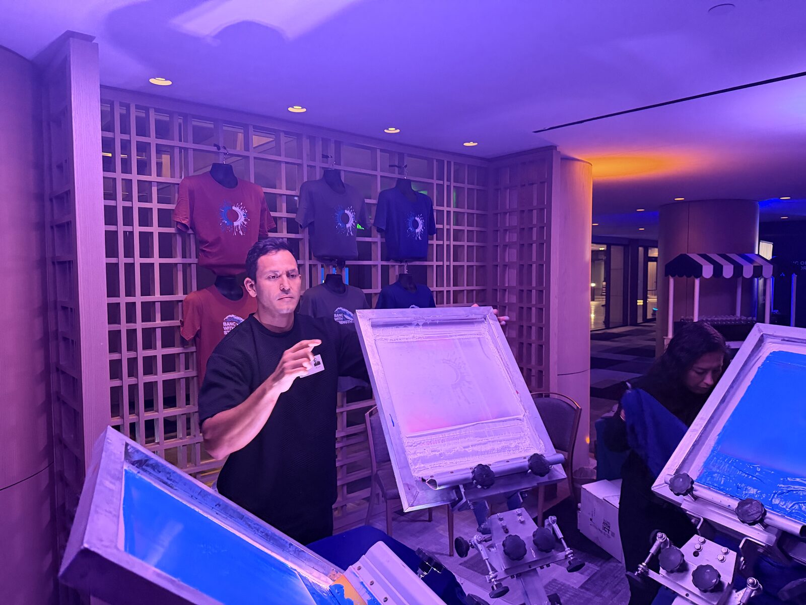 Printer working between two presses with glowing blue screens under purple uplighting beside a wall of display tees
