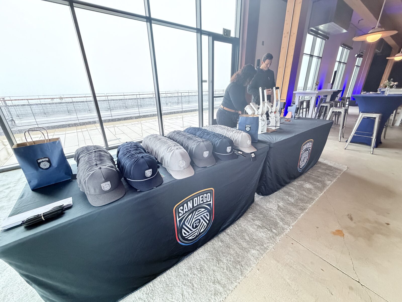 Rows of gray, navy, and cream caps lined up on black San Diego FC table runners in front of floor-to-ceiling windows