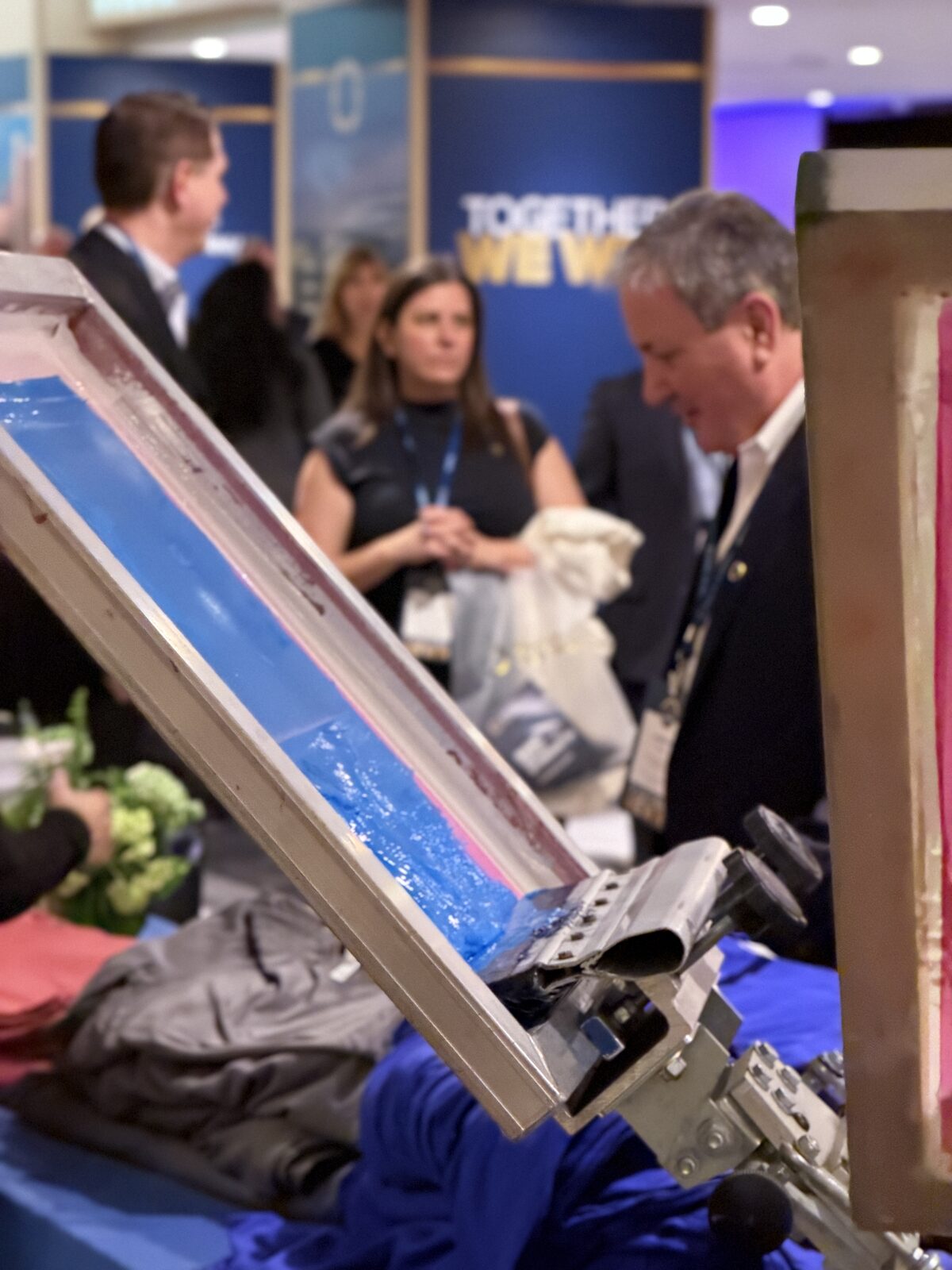 Close-up of a screen printing squeegee mid-stroke through blue ink with event guests blurred behind the frame
