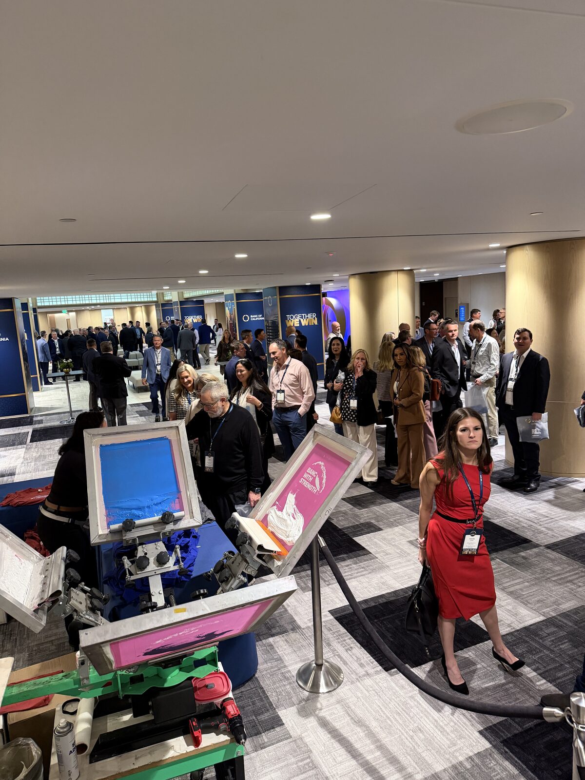 Conference corridor packed with badge-wearing attendees queued past stanchions beside two screen printing presses